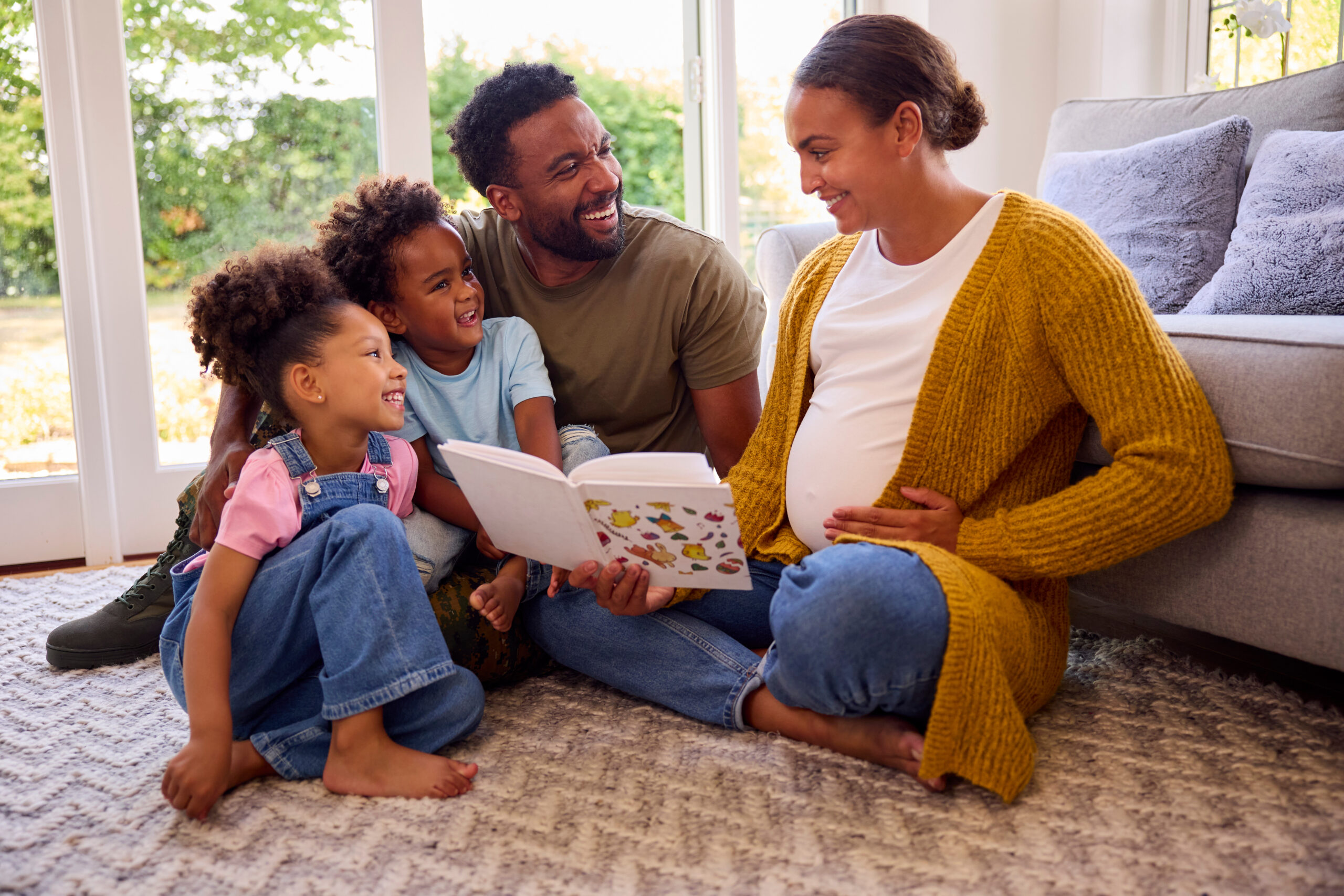 Family With Pregnant Mother Sitting On Floor In Lounge At Home Reading Book Together